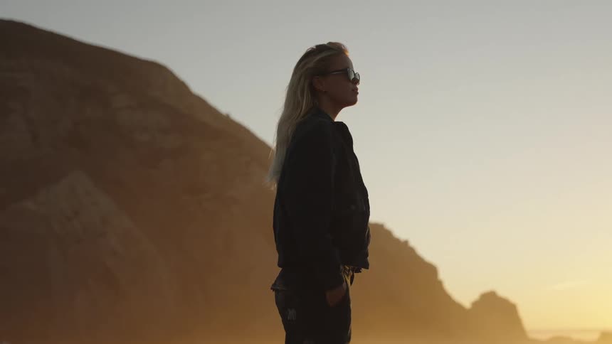 A blonde woman with sunglasses standing on the beach next to cliff with backlight from the sunset
