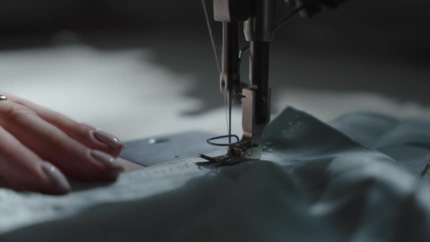A close view shows the needle and presser foot of a sewing machine stitching two pieces of dark fabric together with a persons hands guiding the material in a dimly lit workspace