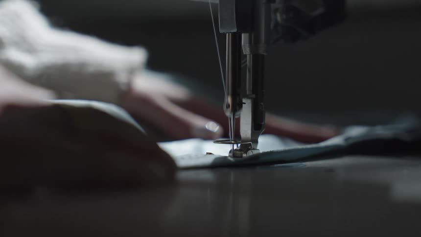 A close view shows the needle and presser foot of a sewing machine stitching two pieces of dark fabric together with a persons hands guiding the material in a dimly lit workspace