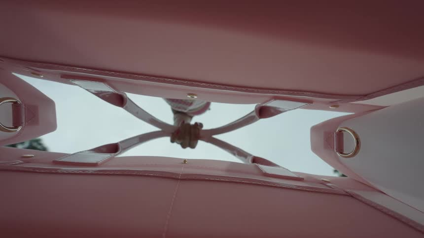 An underside view from within a glossy pink purse or tote bag shows the bags structural support which consists of crisscrossed light pink straps converging in the center in the background a persons hand is holding the bags strap with a finger