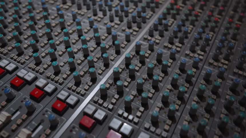 The video is a static closeup shot focusing on the faders and knobs of a professional sound mixing console in a dimly lit setting the console features a complex arrangement of faders red and gray buttons and blue or black knobs suggesting a