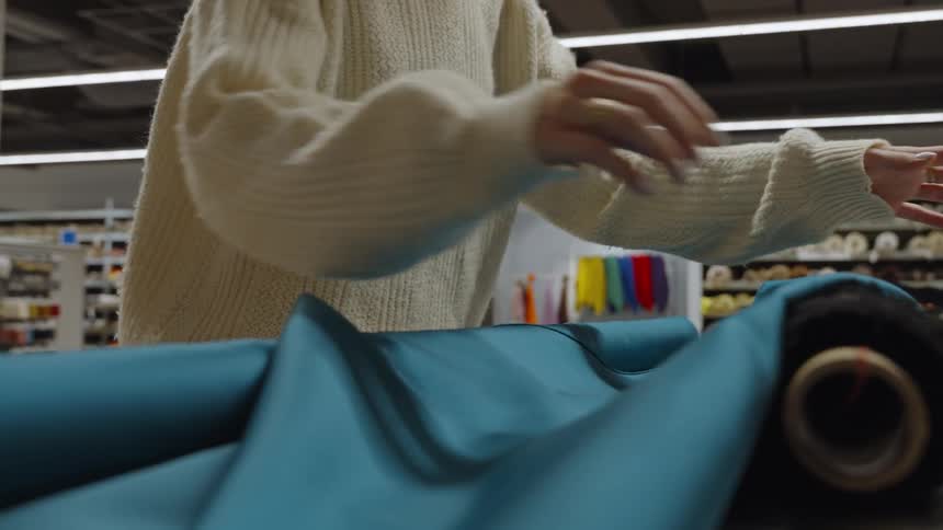 The video is a medium closeup in a warehouse setting showing a persons hands checking the drape of a bolt of vibrant teal fabric they are inspecting the material as it rolls out onto a table from the bolt next to a roll of black fuzzy material