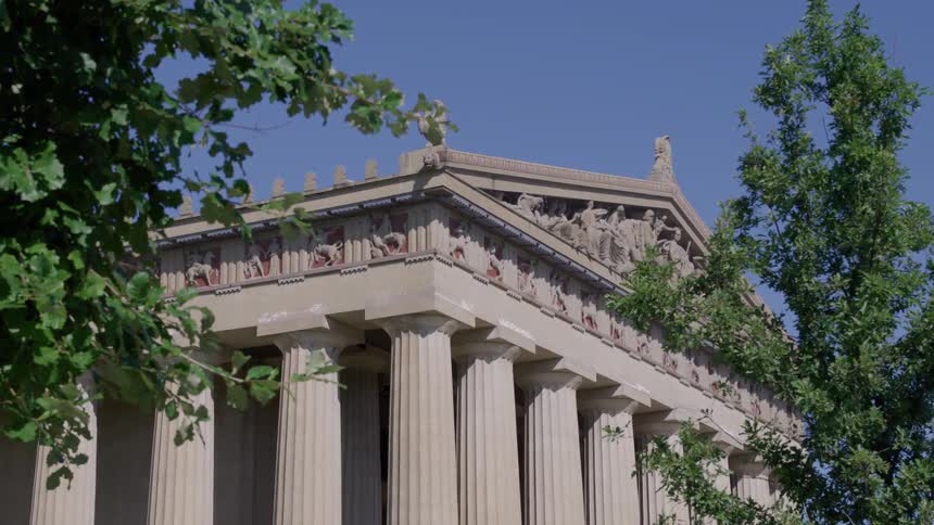 The upper portion of a large classical building tall rows of fluted columns support the roof structure detailed friezes and pedimental sculptures decorate the top section and green tree branches frame the architecture against the clear blue sky