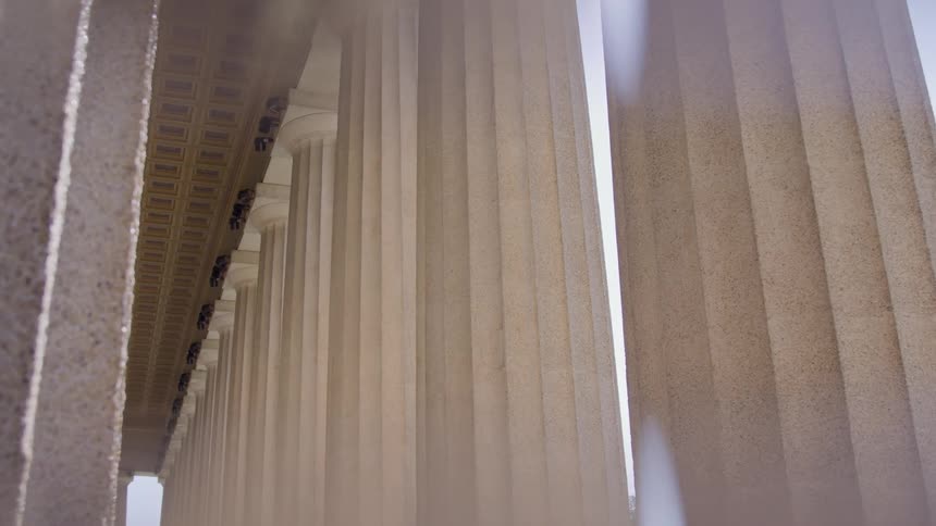 A low angle close view captures a long line of tall cream colored classical columns with decorative friezes along the roofline illuminated by bright sunlight between the two nearest columns