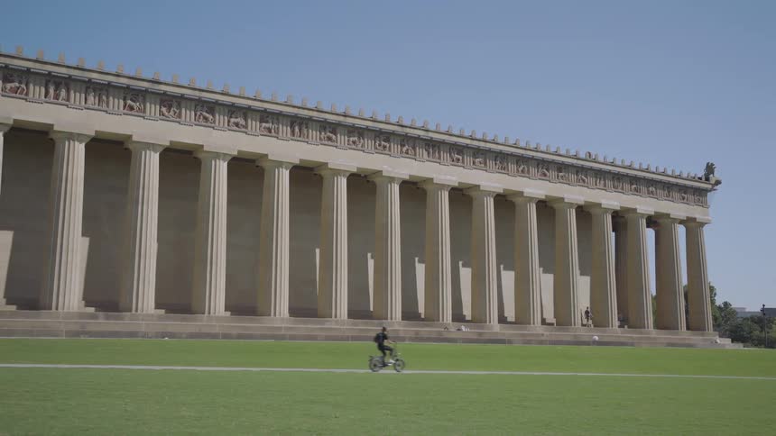 A wide shot shows the exterior of a large classic greek style building with many columns casting long shadows across a green lawn on a sunny day as a person rides a small bicycle in the foreground