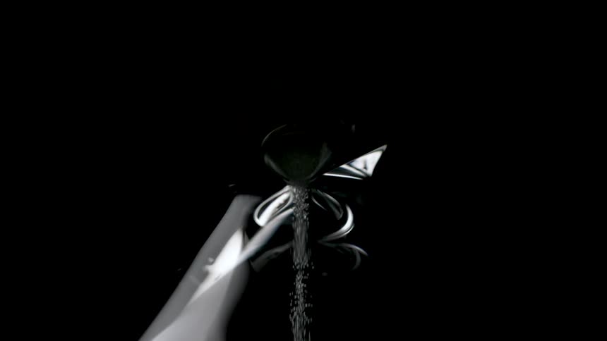 A narrow stream of water pours into a shiny metal sink against a black background
