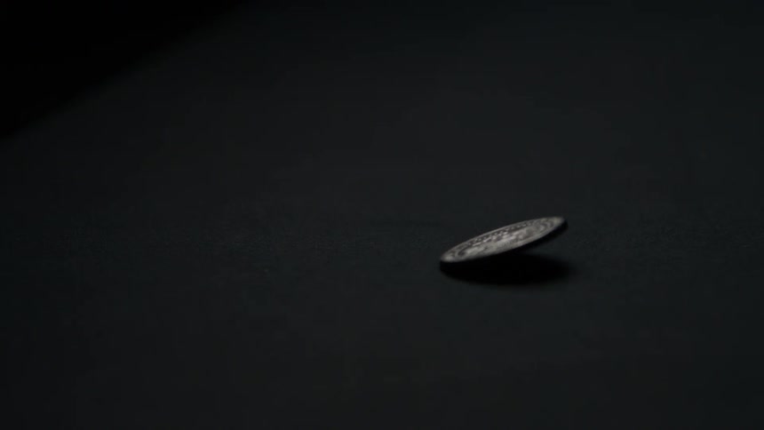 A silver coin spins on its edge the coin wobbles and slows its rotation the coin falls flat onto the surface the surface is a dark solid color and light reflects off the coins metallic faces