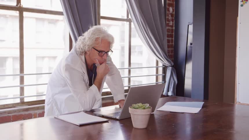 A middle aged male healthcare professional using a laptop computer while leaning on a table in a room during daytime