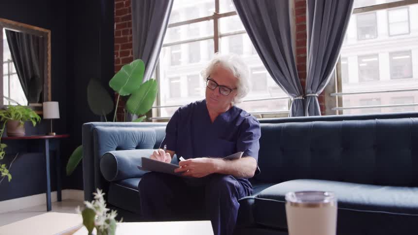 An older man sitting on a couch writing in a notebook with a pen
