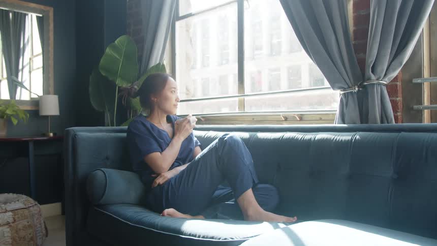 A barefoot woman wearing dark blue pajamas sits alone on a long blue velvet sofa in a moody dimly lit living room sipping from a white mug while gazing out of a large bright window