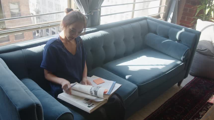 A woman sits on a blue couch reading a textbook in a sunlit room