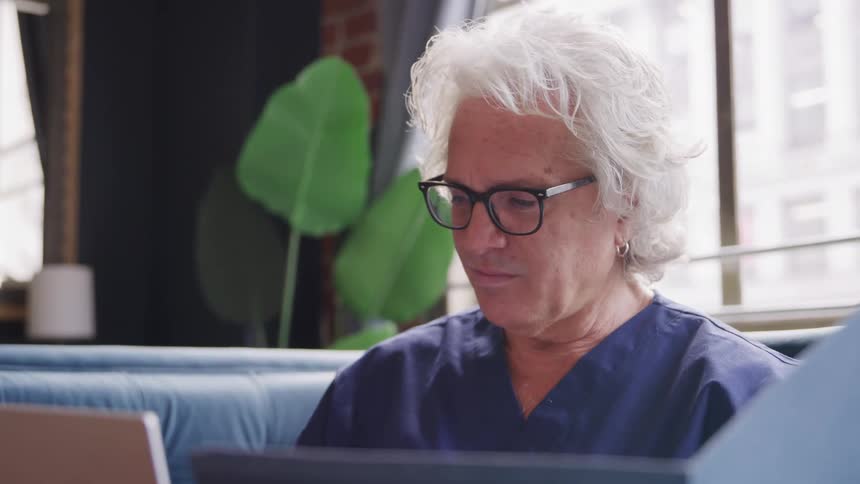 A close view shows an older man with white hair and glasses wearing dark blue medical scrubs looking up from a laptop screen to smile at the camera in a bright modern indoor setting with a plant and large windows