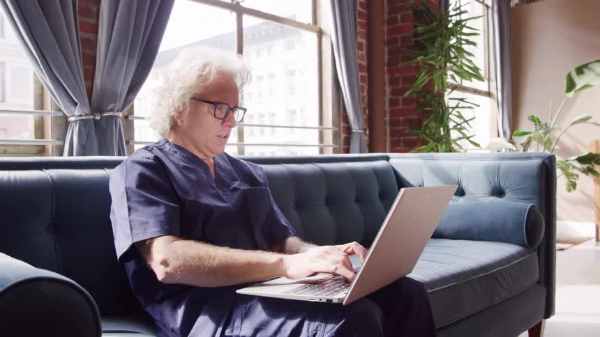 The video shows a middleaged silverhaired man wearing blue medical scrubs sitting on a teal tufted velvet couch in a welllit industrialchic apartment he is concentrating on a silver laptop in his lap periodically pausing to reflect or
