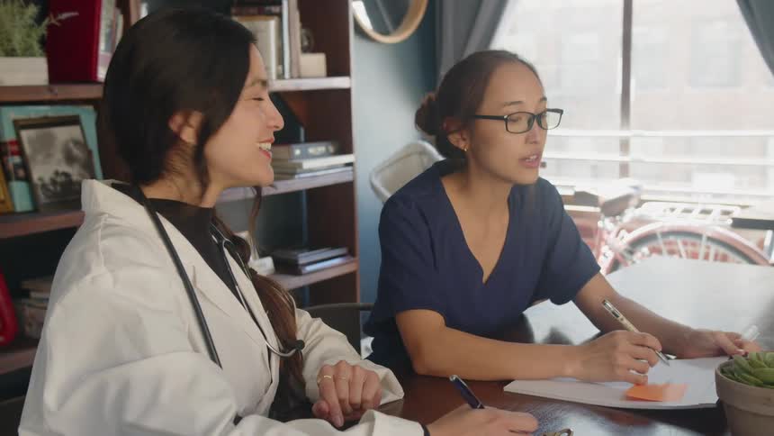 Two women in medical attire sit across a desk from each other one woman wears a white lab coat and smiles while listening the other woman wears dark blue scrubs and talks while pointing at papers and they both hold pens over documents