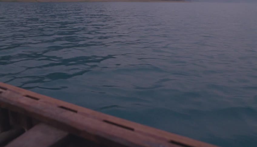 The side of a wooden canoe glides through calm dark blue water creating gentle ripples with a hazy shoreline visible in the distant background