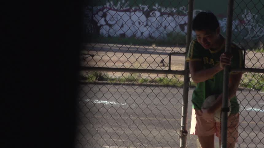 A young man holding a soccer ball, unwinding a chain from a tall chain link fence gate, entering an area, starting to run and dropping the ball