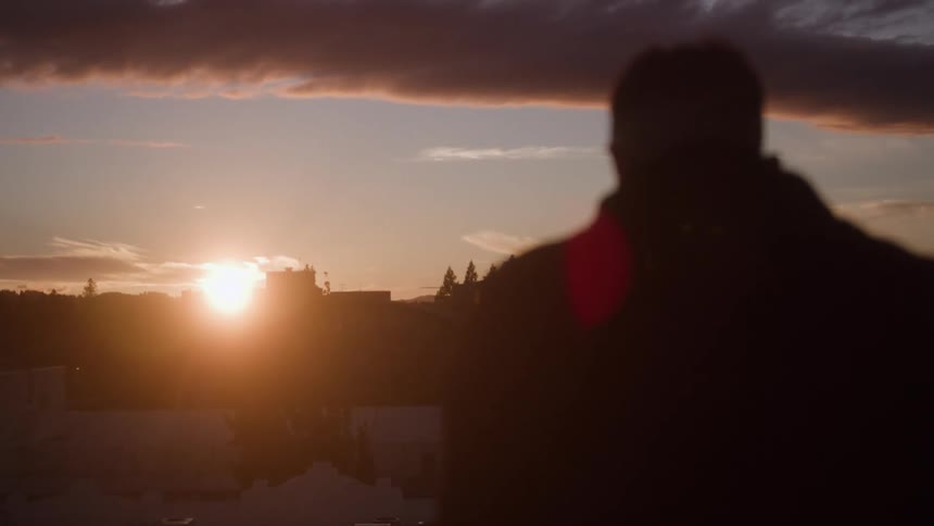 A persons silhouette stands in the foreground looking toward the sunrise on the horizon over a low landscape of buildings and trees all against a sky with dark clouds