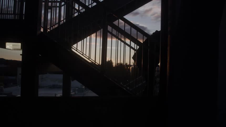 The video shows a silhouetted person descending an outdoor metal staircase at dusk or dawn the structures railing and steps create a dark intersecting pattern against the pale cloudy sky and distant buildings