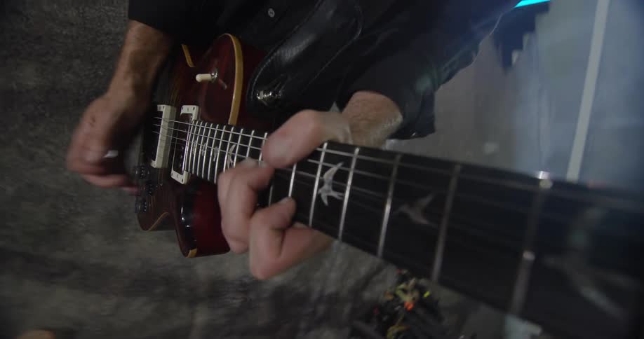 A musicians hands aggressively play a burst finish electric guitar with bird inlays on a stage with a smoky lit background