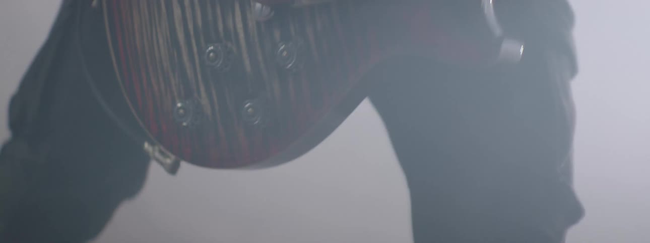 Closeups of a guitars body and a bass guitar are seen partially through a haze of smoke or fog on a dimly lit stage
