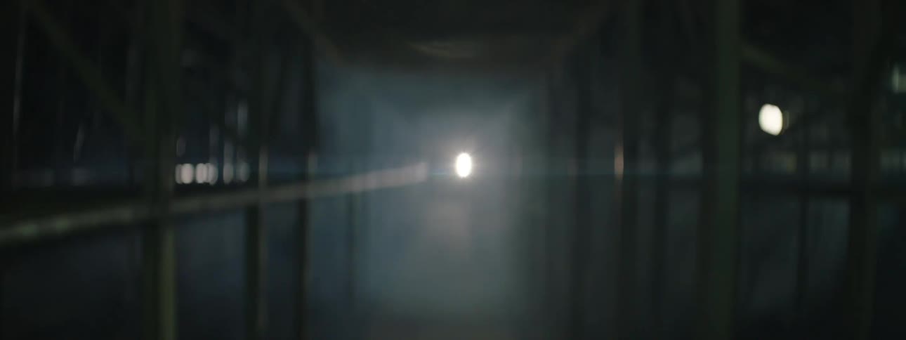 An extreme long view travels through a dark industrial corridor with metal railings and structure as a bright light source shines from the distant center