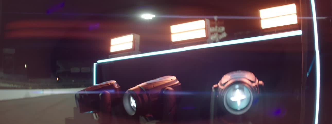 An angled close view of a stage light shows its bright yellow beam and an x shaped aperture with lens flare set against a dark platform illuminated by neon blue strips and rectangular yellow lights