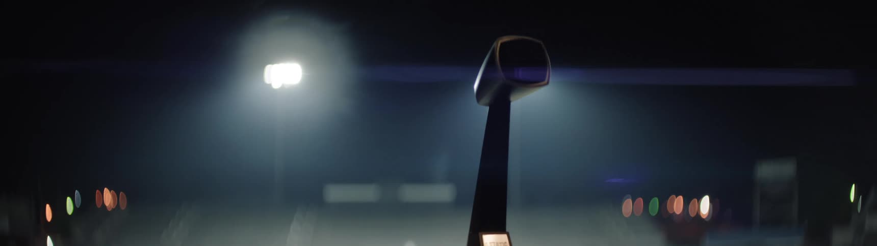 A low angle close view captures a dark microphone on a tall stand illuminated by bright out of focus stadium lights in the dark background which then flicker off leaving the screen black except for small orange lights