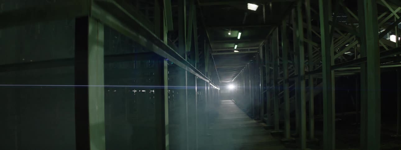 Fluorescent lights flicker over an aisle passing through a structure containing many metal struts