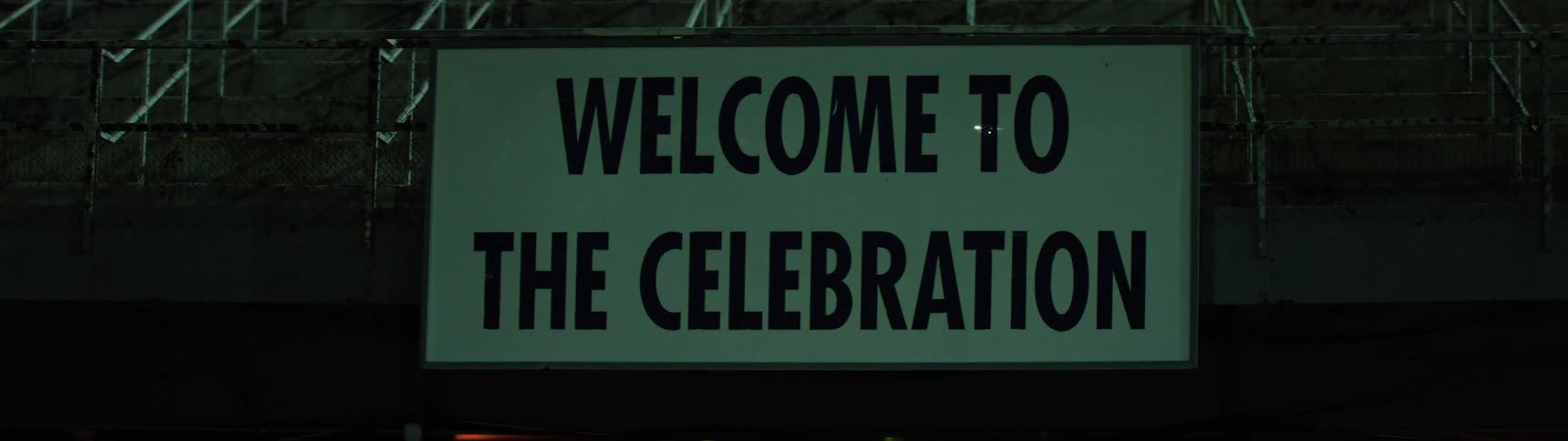 A static shot shows a brightly illuminated sign with the bold black text welcome to the celebration the sign is set against a dark background and light glare is visible on the surface