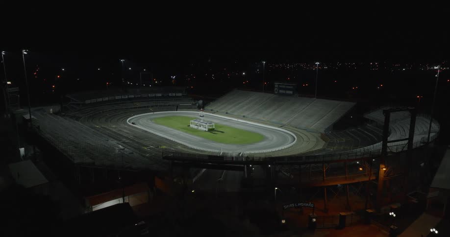 The video is a wide highangle night shot of an empty oval racetrack stadium illuminated by a few distant bright floodlights the camera slowly zooms in on the facility showing the concrete tiered seating and the green inner field of the track