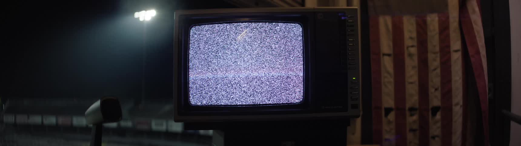 The video is a medium closeup of an old boxy crt television displaying only static on its screen the television is placed outdoors at night with a dimly lit sports or racetrack stadium visible in the blurred background