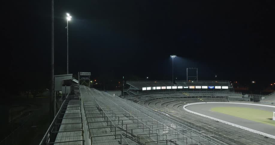 Empty metal bleachers surround oval track lit by floodlights under night sky