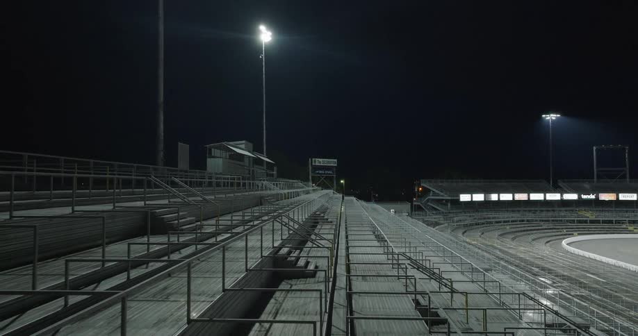 A wide shot captures the long empty rows of metal and concrete stadium bleachers at night a brightly lit floodlight stands tall over the seating area the curve of a racetrack is visible in the lower right
