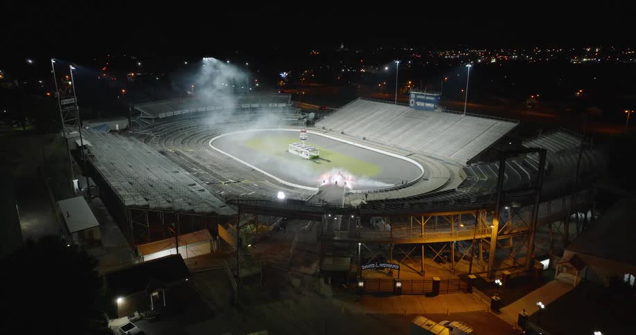 An aerial view shows a large oval racetrack stadium at night bright floodlights illuminate the track surface plumes of mist rise from the center of the circuit and empty tiers of spectator seating surround the dark arena