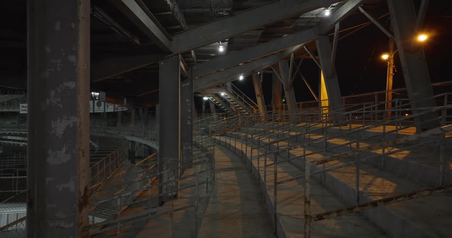 The video shows an interior lowangle shot of the empty illuminated upper seating and structural beams of an old stadium or grandstand at night the area is mostly comprised of steel supports and concrete steps suggesting a rarely used or histo