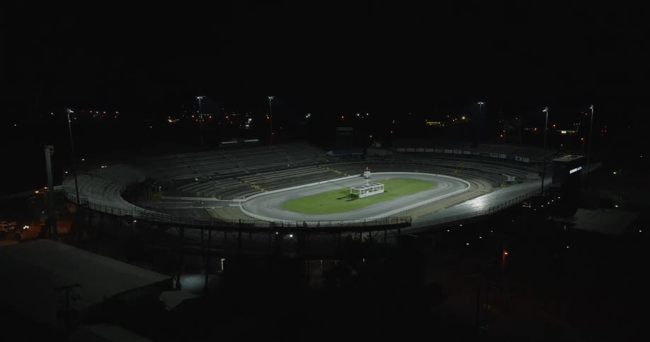 An elevated sweeping aerial shot captures a large empty stadium with bleachers and a race track where a small white structure is visible on the grass infield as the stadium lights are slowly brought up against the dark surrounding city night