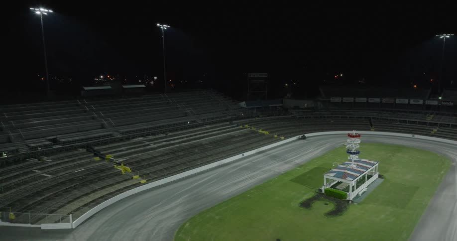An overhead view shows an empty oval racetrack stadium at night a small vehicle drives along the track surface tall lights illuminate the circuit and the rows of dark vacant spectator seating