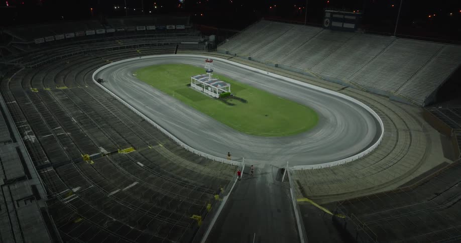 A concrete structure encircles an oval track with a grass center a small building stands on the infield rows of empty spectator seats rise high around the perimeter and the entire area is dark under the night sky