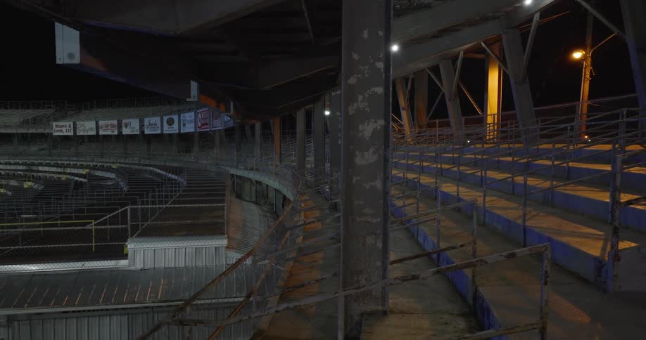 A deserted concrete and metal grandstand sits under harsh night lights and peeling beams