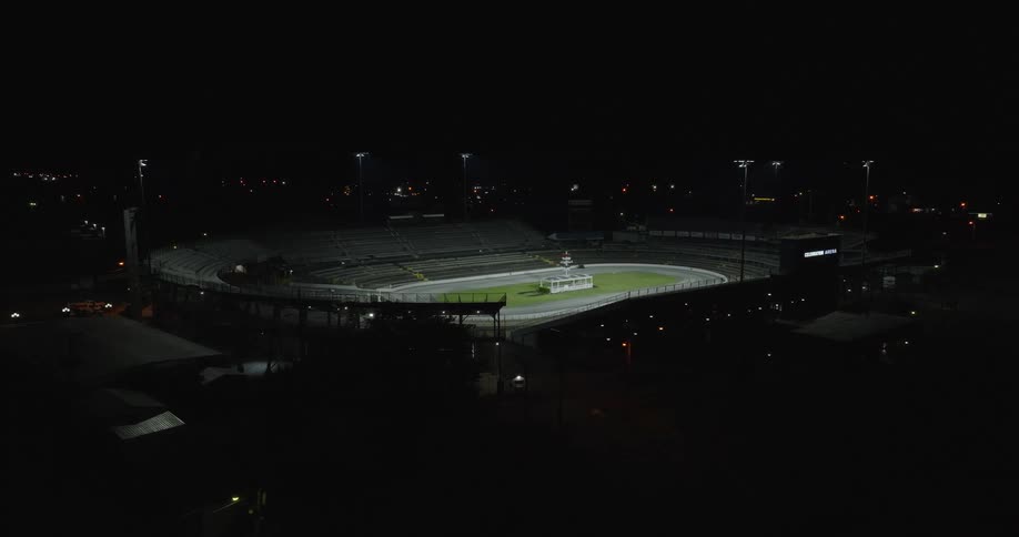 The video is a dark wideangle aerial shot of an empty speedway or dirt track stadium lit up at night the entire stadium is surrounded by darkness and distant scattered lights from a town or city