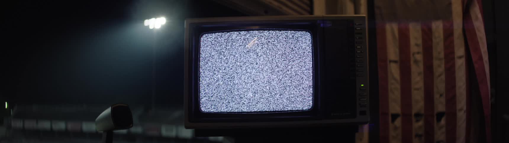 A static shot focuses on the screen of an old dark television monitor the screen displays continuous gray and white static the surrounding area is dark and bright lights are visible outside the frame casting a glow on the left