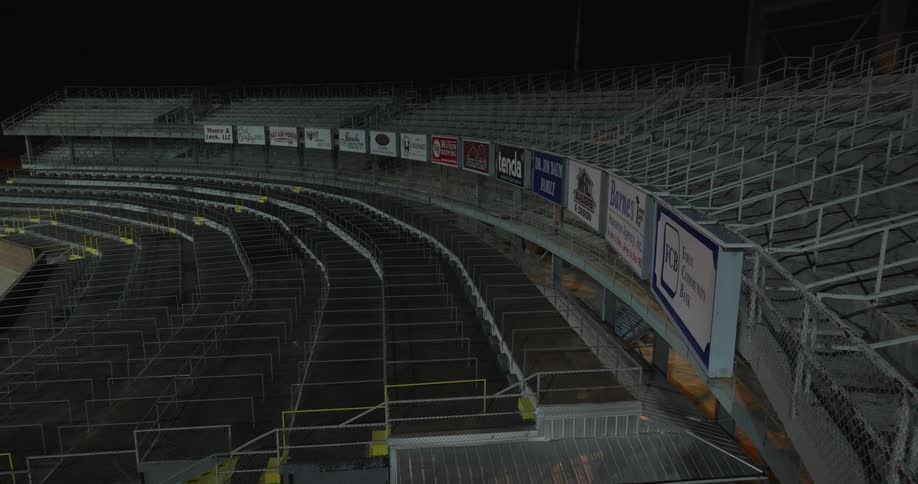 A long wide view of empty curving stadium seating with sponsor signs visible along the railing and exposed metal framework under the dark night sky