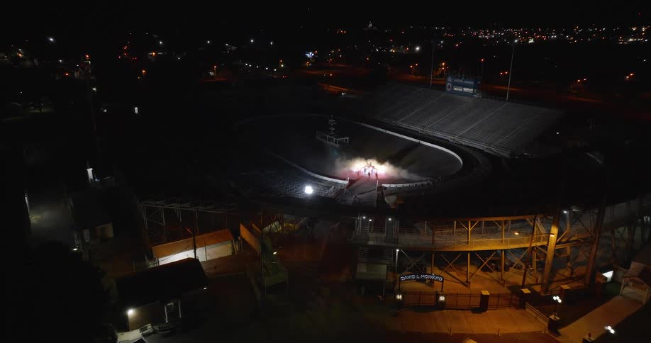 The video is a wide aerial shot of a speedway or dirt track stadium lit up at night the surrounding area is dark with scattered city lights visible in the background