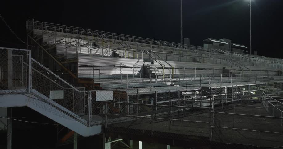 A deserted metal bleacher structure at a stadium glows under bright night lights