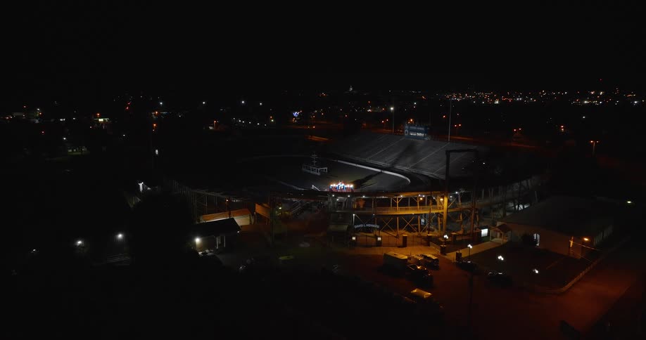 An elevated sweeping aerial shot captures a large empty stadium with bleachers and a race track where a small utility vehicle is visible on the grass infield as the stadium lights are dimmed while bright lights stay flashing in the tracks