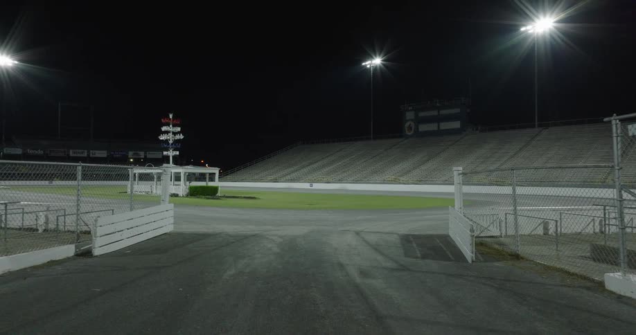 Fenced track entrance with illuminated grandstands in an empty racetrack at night