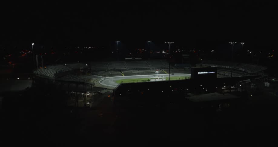 An aerial view shows a largely dark stadium containing an oval track and a grass infield at night tall light towers dimly illuminate the track and tiers of empty seating the building and its surroundings are visible against the dark sky