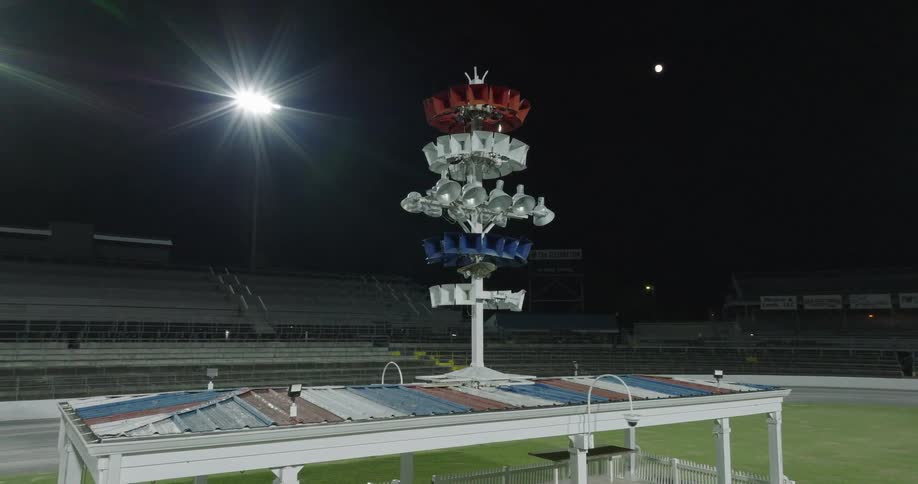 The video features a tall multitiered loudspeaker and lighting assembly with red white and blue elements positioned atop a small structure at a racetrack or stadium the shot is taken at night with the empty grandstands visible in the ba