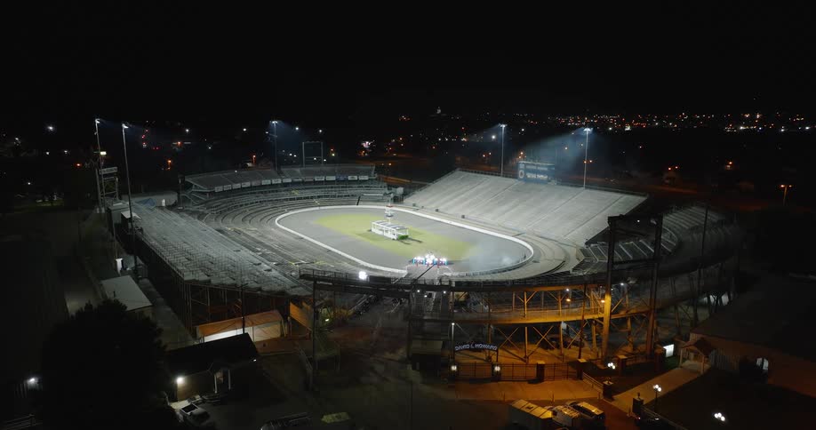 An aerial view shows a brightly lit oval racetrack and surrounding stadium at night rows of empty seating surround the track some vehicles parked on exterior of stadium and distant city lights are visible on the horizon
