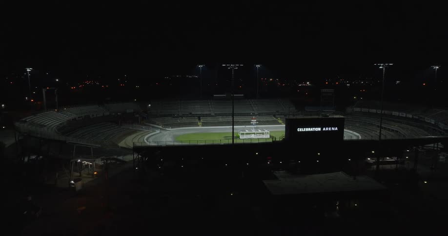 An overhead wide shot shows the floodlights of a stadium slowly turning off at night plunging the oval track and central green field into near darkness a digital display on the right side of the frame still shows the words celebration ar